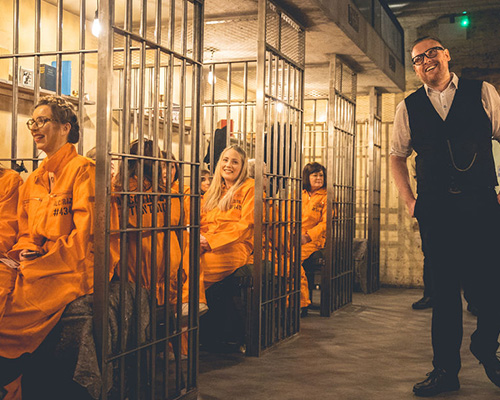The Warden character at Alcotraz immersive experience interacting with customer inmates in prison-themed cocktail bar.